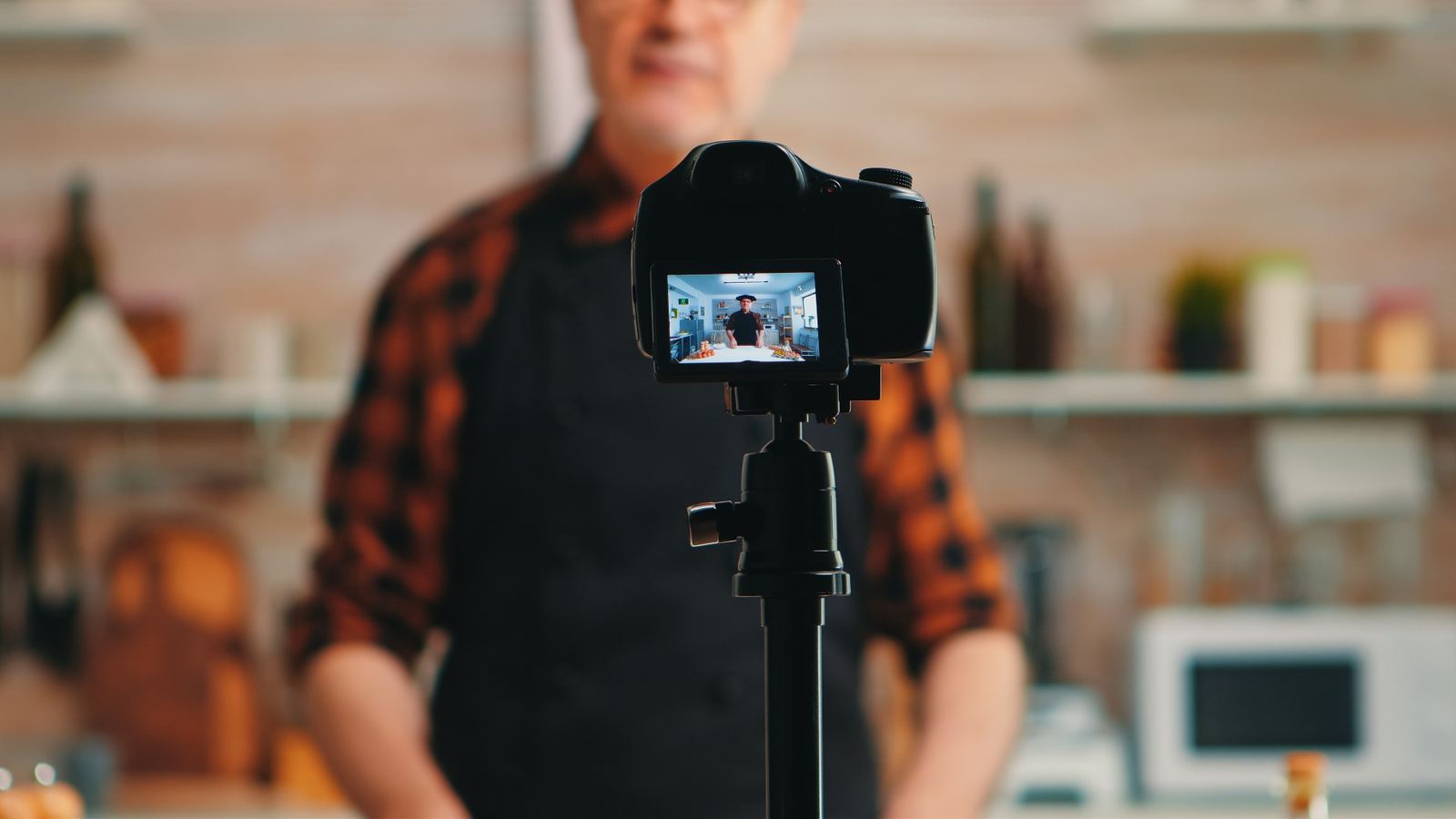 A baker recording a video tutorial to a camera on a tripod in a kitchen.